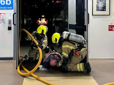 Two firefighters in full breathing apparatus pull a casualty from a dark hospital ward corridor, with a fire hose trailing behind them.