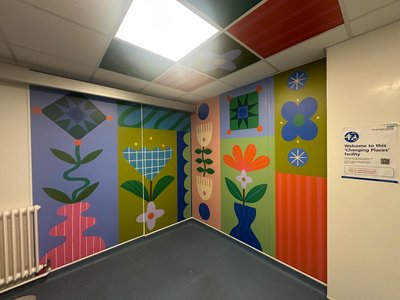 A Changing Places facility in a hospital, featuring vibrant, floor-to-ceiling mural artwork across the walls and ceiling. The colourful design includes stylised flowers, leaves, geometric patterns and abstract botanical shapes in shades of blue, green, pink, orange and yellow.