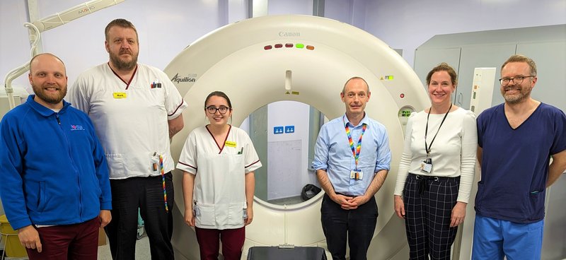 Autocontour software team of 6 people, 2 women and 4 men, stand in front of a CT scanner in a light coloured room.