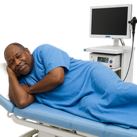 A man in a blue hospital gown lying on a medical bed with a monitor behind him.