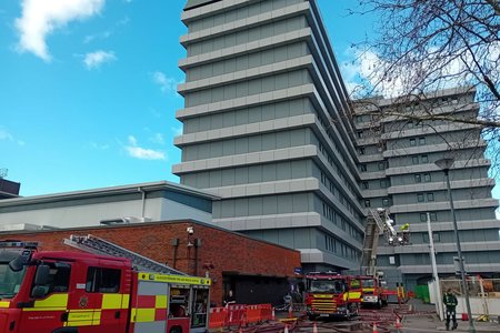 Several fire engines and firefighters are positioned outside a tall hospital building (Gloucestershire Royal Tower Block), with hoses laid out on the ground during an emergency response drill or incident.