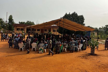 Large group of people gathered under a canopy outside a low building, with chairs arranged in rows