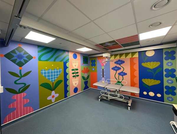A Changing Places facility in a hospital, featuring vibrant, floor-to-ceiling mural artwork across the walls and ceiling. The colourful design includes stylised flowers, leaves, geometric patterns and abstract botanical shapes in shades of blue, green, pink, orange and yellow.