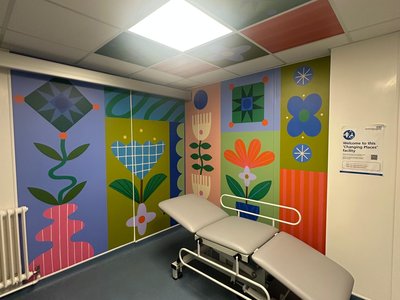 A Changing Places facility in a hospital, featuring vibrant, floor-to-ceiling mural artwork across the walls and ceiling. The colourful design includes stylised flowers, leaves, geometric patterns and abstract botanical shapes in shades of blue, green, pink, orange and yellow.