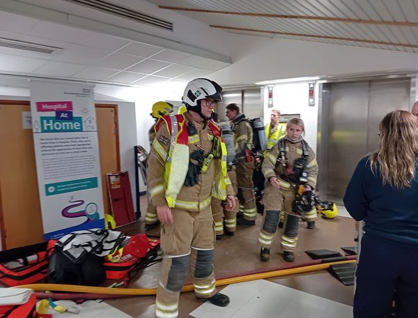 Firefighters in full protective gear gather inside a hospital corridor near a lift lobby, preparing equipment and hoses as part of an emergency response exercise. A “Hospital at Home” information stand and medical bags are visible in the background.